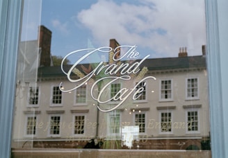 A glass window with elegant calligraphy advertising 'The Grand Café.' The reflection in the window shows a row of historic buildings and a partly cloudy sky. Below the main text, there is a tagline that reads 'The first coffee house in England, in the year 1650.'