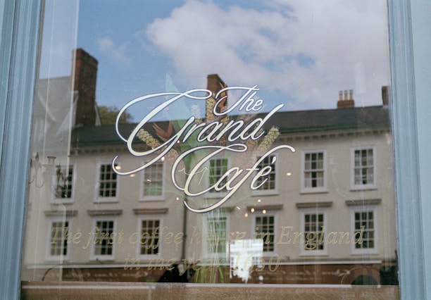 A glass window with elegant calligraphy advertising 'The Grand Café.' The reflection in the window shows a row of historic buildings and a partly cloudy sky. Below the main text, there is a tagline that reads 'The first coffee house in England, in the year 1650.'