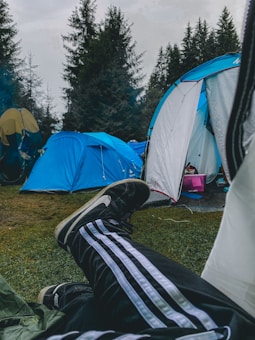 Camping scene featuring blue tents and a person lounging with feet visible, wearing striped track pants and Nike shoes. The campsite is surrounded by tall pine trees and the ground is covered with grass.