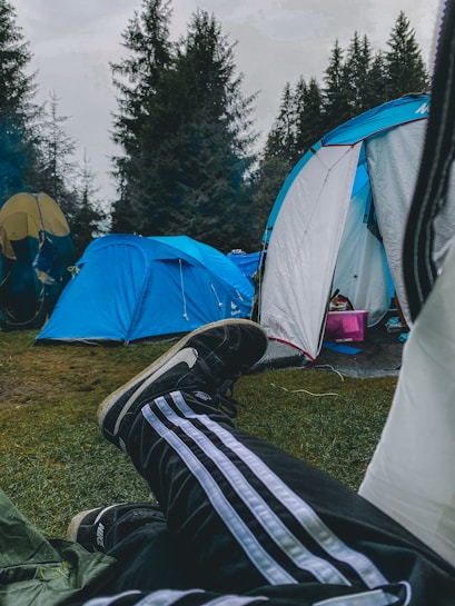 Camping scene featuring blue tents and a person lounging with feet visible, wearing striped track pants and Nike shoes. The campsite is surrounded by tall pine trees and the ground is covered with grass.