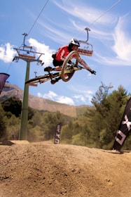 A person riding a mountain bike mid-air against a backdrop of a bright blue sky and rugged terrain. The rider, wearing a red jersey and helmet, is performing a jump with their bike angled diagonally. Lift cables are visible above, alongside wooded hillsides.