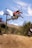 A person riding a mountain bike mid-air against a backdrop of a bright blue sky and rugged terrain. The rider, wearing a red jersey and helmet, is performing a jump with their bike angled diagonally. Lift cables are visible above, alongside wooded hillsides.