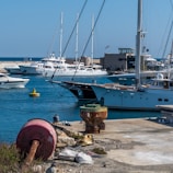 A marina with several yachts and sailboats docked at the harbor. The turquoise waters are calm, and the sky is clear and blue. There are various maritime objects on the dock, including a large rusted buoy and some bags. A person is sitting near the water, appearing relaxed and contemplative.