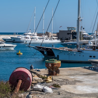 A marina with several yachts and sailboats docked at the harbor. The turquoise waters are calm, and the sky is clear and blue. There are various maritime objects on the dock, including a large rusted buoy and some bags. A person is sitting near the water, appearing relaxed and contemplative.