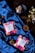 Two colorful tea packages labeled 'TEACORA' are placed on a luxurious blue satin fabric. Nearby, there are two pine cones and a tea bag in a black dish. The scene is set on a dark wooden surface, creating a contrast with the vibrant colors of the products.