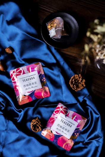 Two colorful tea packages labeled 'TEACORA' are placed on a luxurious blue satin fabric. Nearby, there are two pine cones and a tea bag in a black dish. The scene is set on a dark wooden surface, creating a contrast with the vibrant colors of the products.