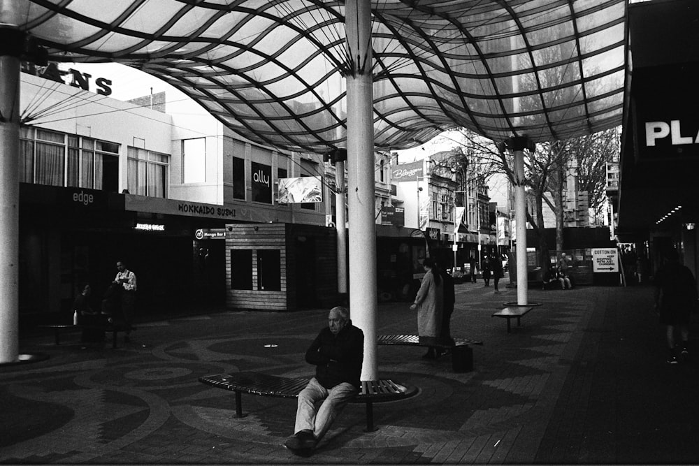 grayscale photo of man sitting on bench near post lamp