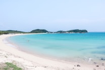 A serene beach scene with turquoise waters stretching into the distance, bordered by soft white sand. Small groups of people are leisurely enjoying the water near the shore. Distant green hills provide a lush backdrop, and the clear sky enhances the calm atmosphere.