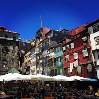 A vibrant street scene with colorful, historic buildings featuring red, yellow, and blue facades. The area is bustling with people sitting at tables under large white umbrellas, indicating a lively caf&eacute; setting. The architecture showcases intricate details and balconies adorned with plants and laundry.