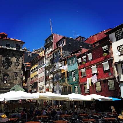 A vibrant street scene with colorful, historic buildings featuring red, yellow, and blue facades. The area is bustling with people sitting at tables under large white umbrellas, indicating a lively caf&eacute; setting. The architecture showcases intricate details and balconies adorned with plants and laundry.