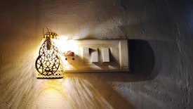 A decorative lamp with intricate patterns casts a warm light on a wall-mounted electrical switch panel. The lamp creates complex shadows on the wall, enhancing the textured surface.