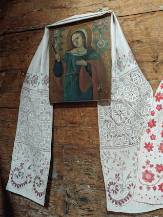 A religious icon painted on wood depicts a haloed figure holding a cross, dressed in traditional garments with intricate patterns. The icon is draped with a decorative cloth featuring elaborate embroidery and floral designs, likely handmade. The background consists of a wooden surface, adding a rustic and historical feel to the scene.