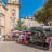 A bustling outdoor market scene with people shopping for flowers and plants under colorful umbrellas. The marketplace is set against a backdrop of historic buildings and a clock tower, with clear blue skies overhead.