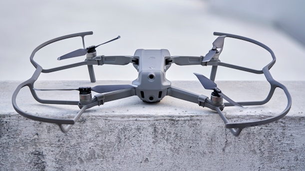 A sleek, modern drone sits on a concrete surface, equipped with a robust frame and four propellers. It appears to be designed for stability and efficiency, with aerodynamic features and a sturdy build. The drone's color seamlessly blends with its industrial setting.