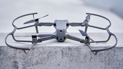 A sleek, modern drone sits on a concrete surface, equipped with a robust frame and four propellers. It appears to be designed for stability and efficiency, with aerodynamic features and a sturdy build. The drone's color seamlessly blends with its industrial setting.