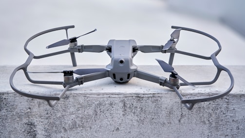 A sleek, modern drone sits on a concrete surface, equipped with a robust frame and four propellers. It appears to be designed for stability and efficiency, with aerodynamic features and a sturdy build. The drone's color seamlessly blends with its industrial setting.