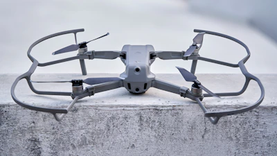 A sleek, modern drone sits on a concrete surface, equipped with a robust frame and four propellers. It appears to be designed for stability and efficiency, with aerodynamic features and a sturdy build. The drone's color seamlessly blends with its industrial setting.
