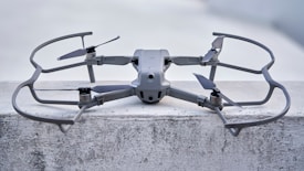 A sleek, modern drone sits on a concrete surface, equipped with a robust frame and four propellers. It appears to be designed for stability and efficiency, with aerodynamic features and a sturdy build. The drone's color seamlessly blends with its industrial setting.