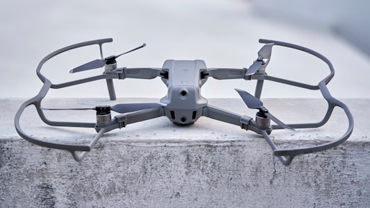 Een gestroomlijnde, moderne drone staat op een betonnen oppervlak, uitgerust met een robuust frame en vier propellers. Hij lijkt ontworpen te zijn voor stabiliteit en efficiëntie, met aerodynamische kenmerken en een stevige constructie. De kleur van de drone past naadloos bij de industriële omgeving.