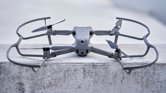 A sleek, modern drone sits on a concrete surface, equipped with a robust frame and four propellers. It appears to be designed for stability and efficiency, with aerodynamic features and a sturdy build. The drone's color seamlessly blends with its industrial setting.