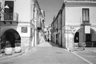 A narrow cobblestone street flanked by historic buildings with arched entrances and balconies. A sign for 'Caffe 41 Piazza Umberto' is visible on the left side. Potted plants line the street, and a few people sit outside the cafe. Several street signs and a parked car can be seen in the distance.