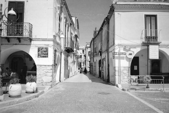 A narrow cobblestone street flanked by historic buildings with arched entrances and balconies. A sign for 'Caffe 41 Piazza Umberto' is visible on the left side. Potted plants line the street, and a few people sit outside the cafe. Several street signs and a parked car can be seen in the distance.