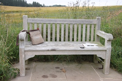 A weathered wooden bench is set against a backdrop of lush green fields and a distant tree line. On the bench, there is a beige shoulder bag resting on the left armrest and a white hardcover book with a pair of sunglasses beside it placed on the seat. The words 'In Memory of Jenny Nelson A True Country Woman' are engraved on the bench.