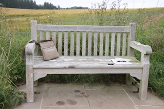 A weathered wooden bench is set against a backdrop of lush green fields and a distant tree line. On the bench, there is a beige shoulder bag resting on the left armrest and a white hardcover book with a pair of sunglasses beside it placed on the seat. The words 'In Memory of Jenny Nelson A True Country Woman' are engraved on the bench.