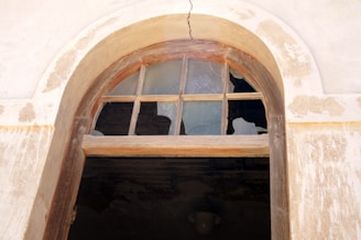 An old, arched window with broken glass panes and a wooden frame is set into a weathered, beige wall. Cracks and peeling paint are visible around the window, suggesting deterioration over time.