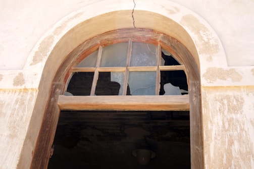 An old, arched window with broken glass panes and a wooden frame is set into a weathered, beige wall. Cracks and peeling paint are visible around the window, suggesting deterioration over time.