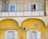 A person standing on a second-floor balcony of a yellow building, peering over a railing. Below, a basket is suspended by a string against the backdrop of the building's facade. The architecture includes arched features, and there's an air conditioning unit and antenna affixed to the wall.