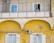 A person standing on a second-floor balcony of a yellow building, peering over a railing. Below, a basket is suspended by a string against the backdrop of the building's facade. The architecture includes arched features, and there's an air conditioning unit and antenna affixed to the wall.