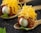 A gourmet dish is artfully presented on a rustic spoon. The dish features a small, glossy white egg encased in a layer of thinly sliced cured meat. On top, a generous amount of finely shredded yellow cheese adds texture and color. The spoon rests on a dark, textured surface, with a small green leaf placed nearby for garnish, contributing to the overall elegance of the presentation.
