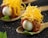 A gourmet dish is artfully presented on a rustic spoon. The dish features a small, glossy white egg encased in a layer of thinly sliced cured meat. On top, a generous amount of finely shredded yellow cheese adds texture and color. The spoon rests on a dark, textured surface, with a small green leaf placed nearby for garnish, contributing to the overall elegance of the presentation.