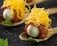 A gourmet dish is artfully presented on a rustic spoon. The dish features a small, glossy white egg encased in a layer of thinly sliced cured meat. On top, a generous amount of finely shredded yellow cheese adds texture and color. The spoon rests on a dark, textured surface, with a small green leaf placed nearby for garnish, contributing to the overall elegance of the presentation.