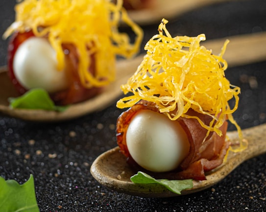 A gourmet dish is artfully presented on a rustic spoon. The dish features a small, glossy white egg encased in a layer of thinly sliced cured meat. On top, a generous amount of finely shredded yellow cheese adds texture and color. The spoon rests on a dark, textured surface, with a small green leaf placed nearby for garnish, contributing to the overall elegance of the presentation.