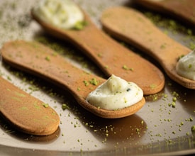 Edible spoons made of a light brown cookie-like material are topped with a dollop of cream or frosting. Green sprinkles, possibly matcha or pistachio, are scattered across the spoons and the reflective surface beneath them, adding a touch of color.