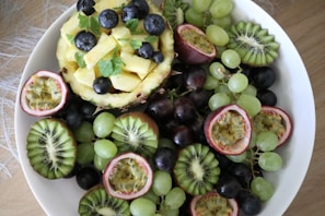 A vibrant assortment of fresh fruits arranged in a white bowl. Slices of kiwi, halved passion fruits, and clusters of green and purple grapes surround a hollowed pineapple filled with pineapple chunks and blueberries. Fresh mint leaves are scattered for added freshness.