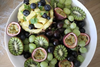 A vibrant assortment of fresh fruits arranged in a white bowl. Slices of kiwi, halved passion fruits, and clusters of green and purple grapes surround a hollowed pineapple filled with pineapple chunks and blueberries. Fresh mint leaves are scattered for added freshness.