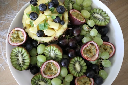 A vibrant assortment of fresh fruits arranged in a white bowl. Slices of kiwi, halved passion fruits, and clusters of green and purple grapes surround a hollowed pineapple filled with pineapple chunks and blueberries. Fresh mint leaves are scattered for added freshness.