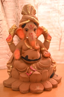 A clay sculpture of the Hindu deity Ganesha, depicted in a seated position. The figure is adorned with gold ornaments and intricate detailing on the headgear and is colored predominantly in earthy tones with accents of pink and black.