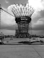 A large metal framework structure is under construction, surrounded by scaffolding and cranes on a wide concrete platform. Overcast skies cast a grey mood over the scene, with heavy clouds scattered throughout.