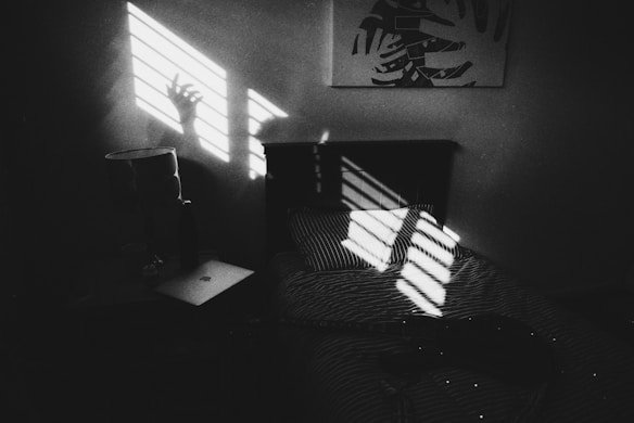 A dimly lit room features a bed with striped sheets, on which a guitar rests. Sunlight streams through blinds, casting striped shadows on the wall and bed. A hand silhouette is visible within the shadows on the wall. A side table next to the bed holds a lamp and a closed laptop. A piece of abstract art hangs on the wall.