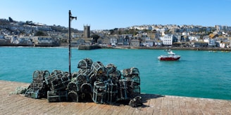 A picturesque coastal town with charming houses spread across a hillside. The foreground features a stack of lobster pots on a pier, set against the backdrop of turquoise waters. A red and white fishing boat sails gracefully in the harbor under a clear blue sky.