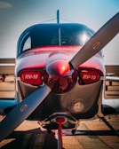 A sleek, modern small aircraft with a red and silver exterior is parked on a runway. The nose is prominently featuring a large propeller, with the designation 'RV-10' clearly visible on both sides of the fuselage. The cockpit canopy is reflective, showing the bright outdoor setting.