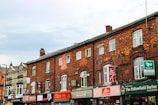 A row of old brick buildings with various small businesses, including restaurants and a barber shop, located on the ground floor. The architecture features arched windows and a mixture of brick and stone materials. Signage for different shops is prominently displayed.