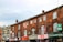 A row of old brick buildings with various small businesses, including restaurants and a barber shop, located on the ground floor. The architecture features arched windows and a mixture of brick and stone materials. Signage for different shops is prominently displayed.