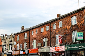 A row of old brick buildings with various small businesses, including restaurants and a barber shop, located on the ground floor. The architecture features arched windows and a mixture of brick and stone materials. Signage for different shops is prominently displayed.