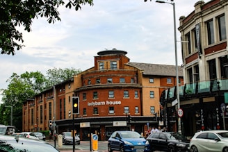 A city street scene featuring 'ladybarn house,' a red-brick building with a curved facade. Several cars are parked and in motion along the street, and there are traffic lights and road signs. The surrounding area includes trees and a partially visible modern glass-fronted building. A few pedestrians are walking on the sidewalk.