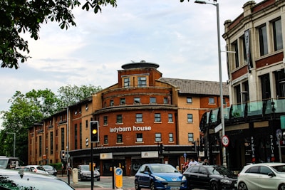 A city street scene featuring 'ladybarn house,' a red-brick building with a curved facade. Several cars are parked and in motion along the street, and there are traffic lights and road signs. The surrounding area includes trees and a partially visible modern glass-fronted building. A few pedestrians are walking on the sidewalk.